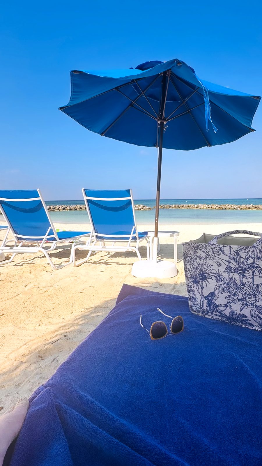 A blue umbrella, two blue loungers and a beach bag on the sand at Sunset Cove, with the rock breakwater behind