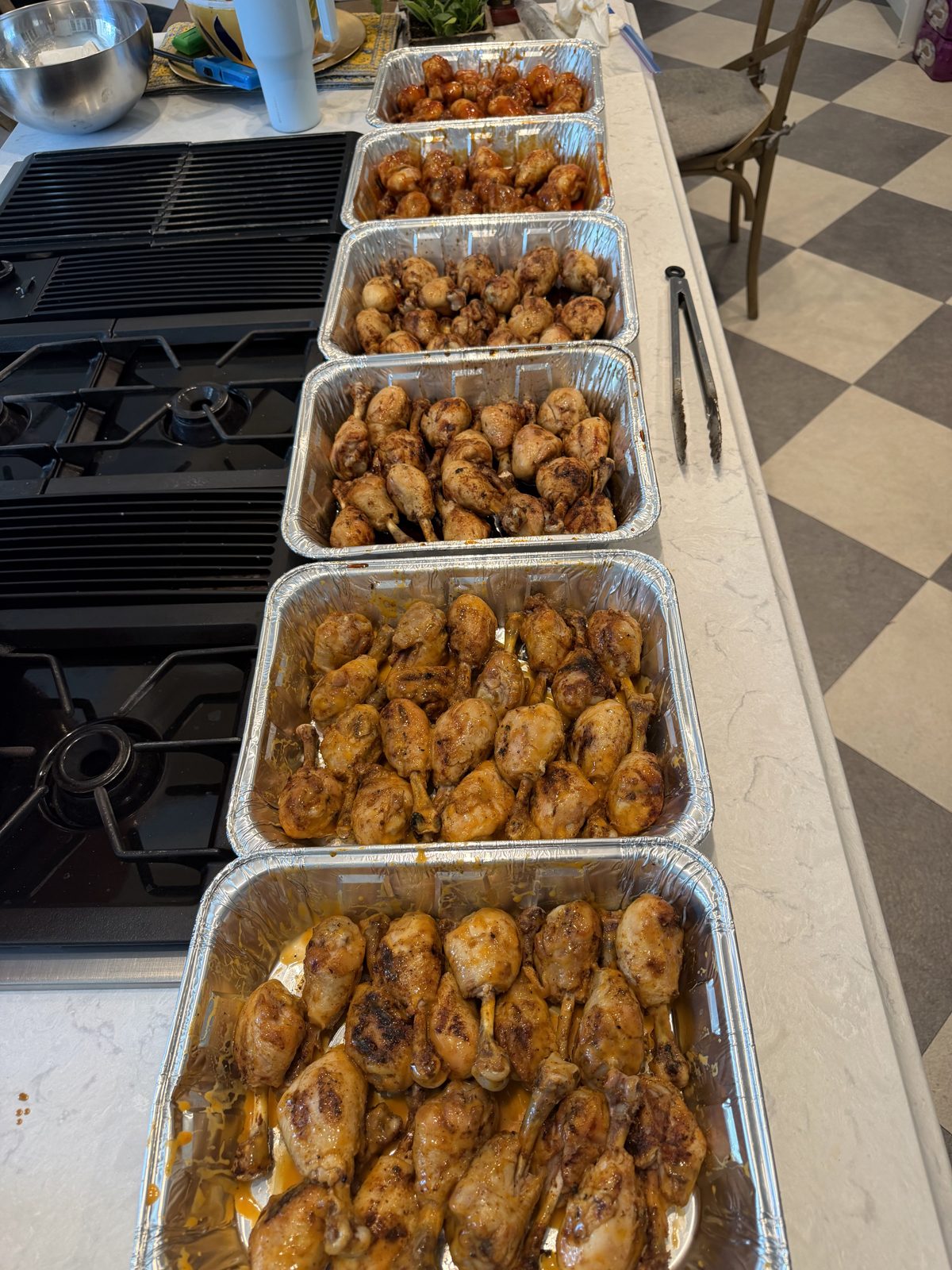 Six foil trays of grilled, sauced drumsticks lined up along a kitchen counter