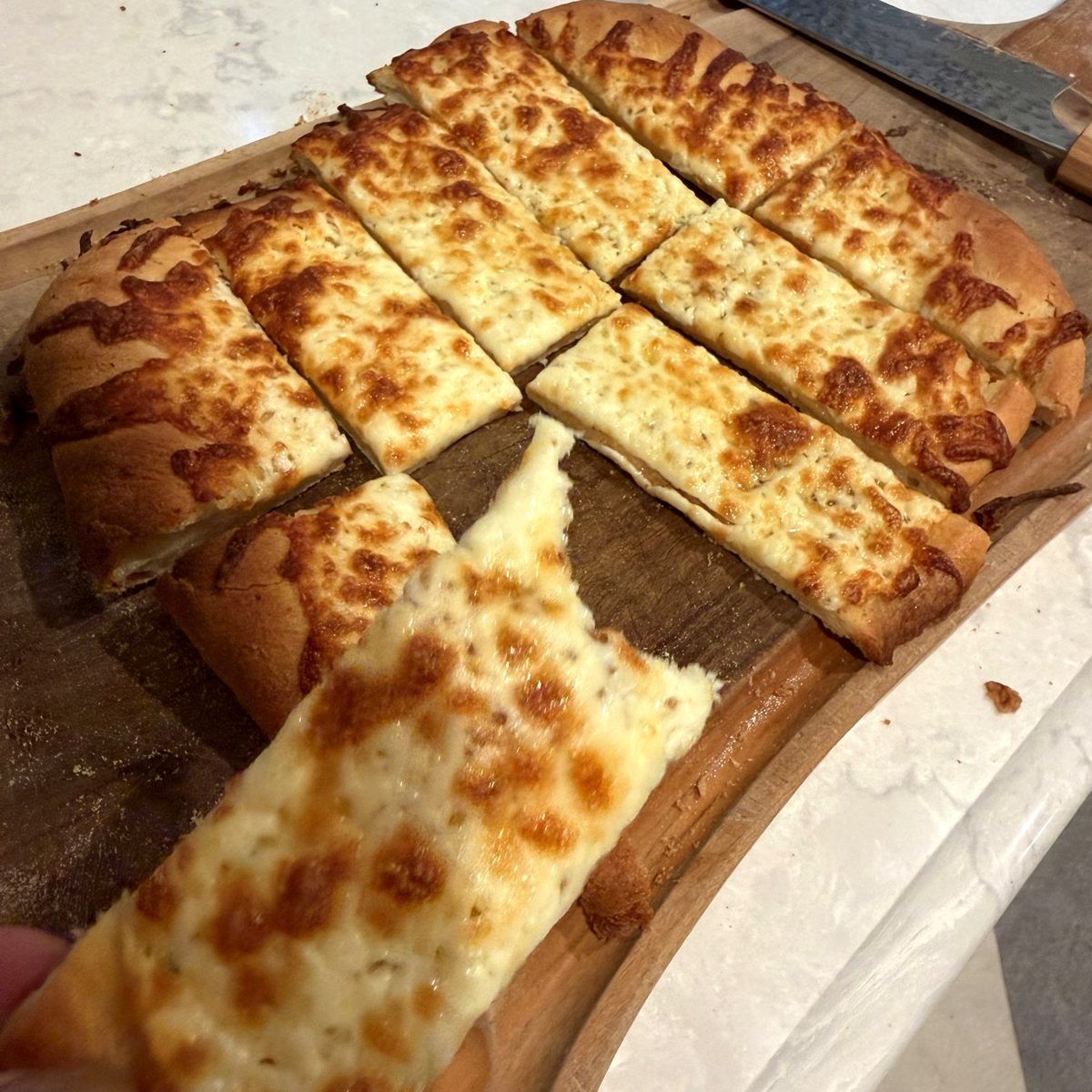 Garlic cheese fingers with browned, bubbling mozzarella cut into strips on a wooden board, one lifted away