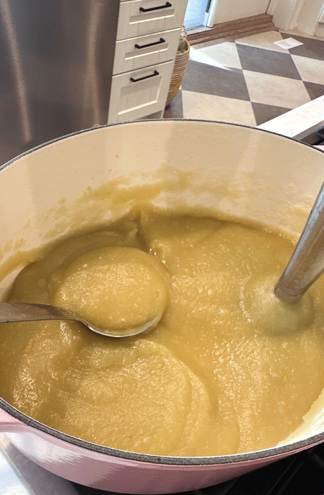 A pink Dutch oven of smooth homemade applesauce with a ladle and an immersion blender
