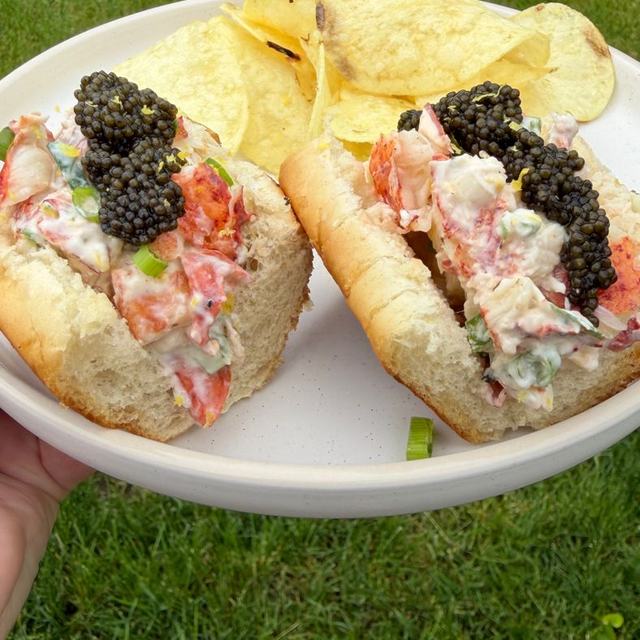 Two East Coast lobster rolls piled with crème fraîche–dressed lobster and topped with caviar, on a plate with potato chips, held over green grass