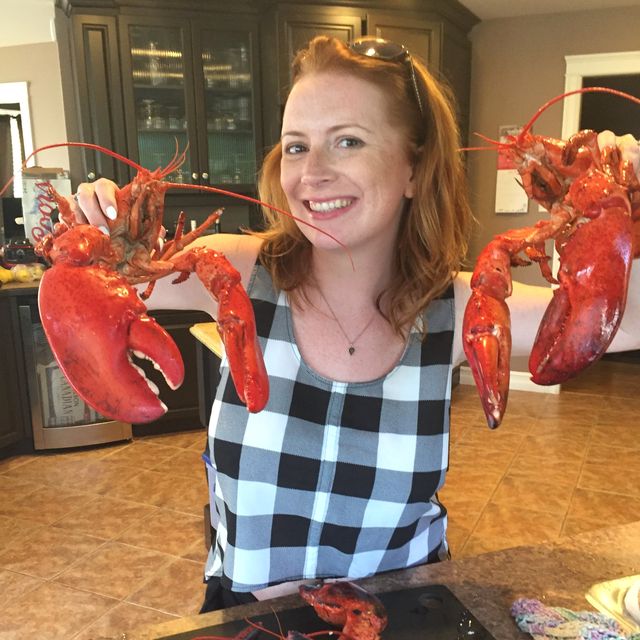 Heather grinning in a kitchen, holding up two cooked lobsters by the claws