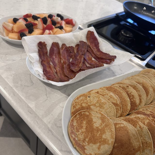 A platter of oatmeal pancakes next to fruit salad and bacon on a kitchen counter