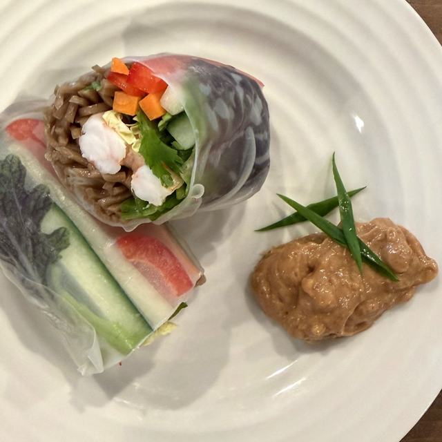 A peanut soba and shrimp fresh roll cut in half to show the cross-section, on a white plate with a dollop of peanut dipping sauce