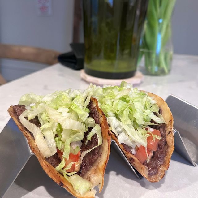 Two crispy smash burger tacos with shredded lettuce and tomato in a steel taco stand