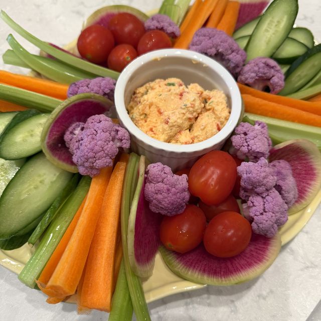 A ramekin of whipped pimento ricotta dip in the middle of a platter of carrots, celery, cucumber, cherry tomatoes, watermelon radish and purple cauliflower