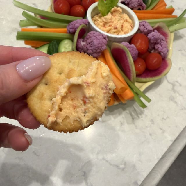 A Ritz cracker loaded with pimento ricotta dip, held over the vegetable platter