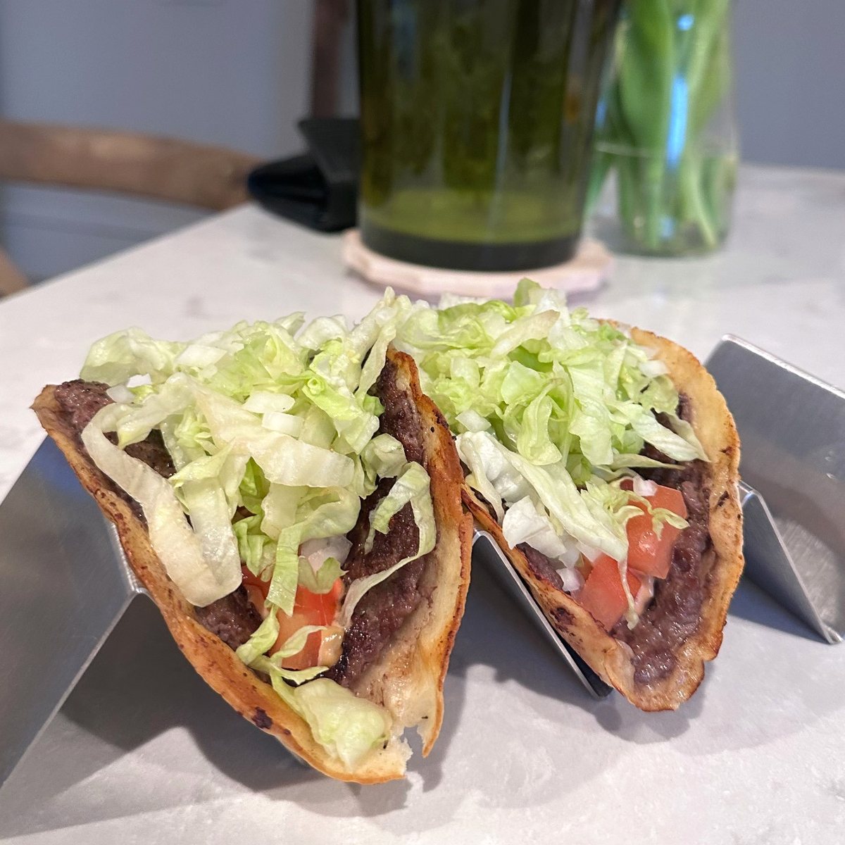 Two crispy smash burger tacos with shredded lettuce and tomato in a steel taco stand
